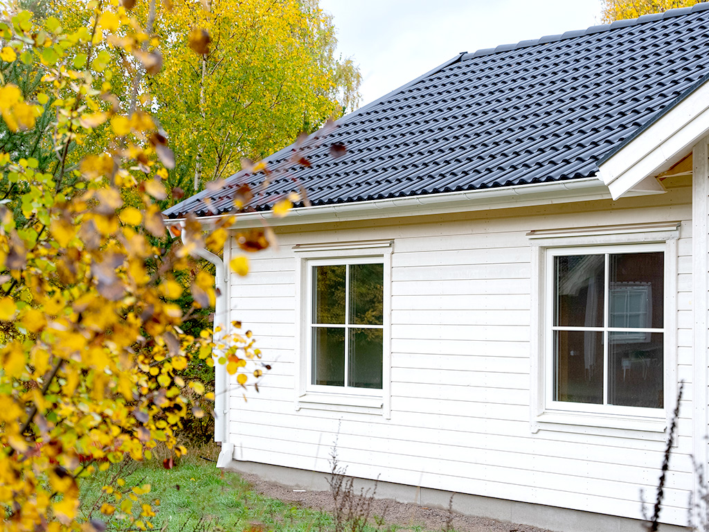 Vit enplansvilla med spröjsade fönster och svart tak, omgiven av höstlöv och grön gräsmatta.
